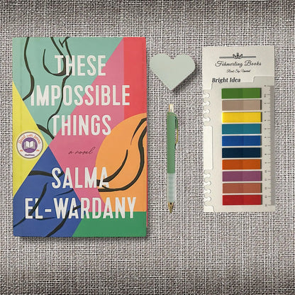 A styled flat-lay featuring the book cover of 'These Impossible Things' by Salma El-Wardany, a pen, a heart shaped leather bookmark, and Bright Idea annotation tabs from Fehmerling Books, all arranged on a textured gray fabric background.