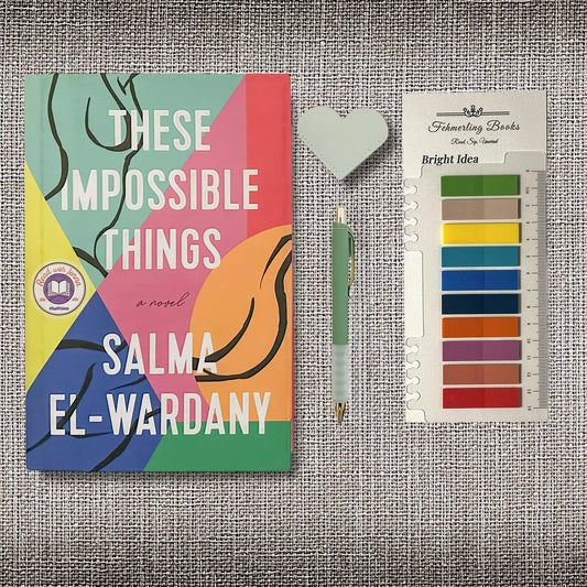 A styled flat-lay featuring the book cover of 'These Impossible Things' by Salma El-Wardany, a pen, a heart shaped leather bookmark, and Bright Idea annotation tabs from Fehmerling Books, all arranged on a textured gray fabric background.