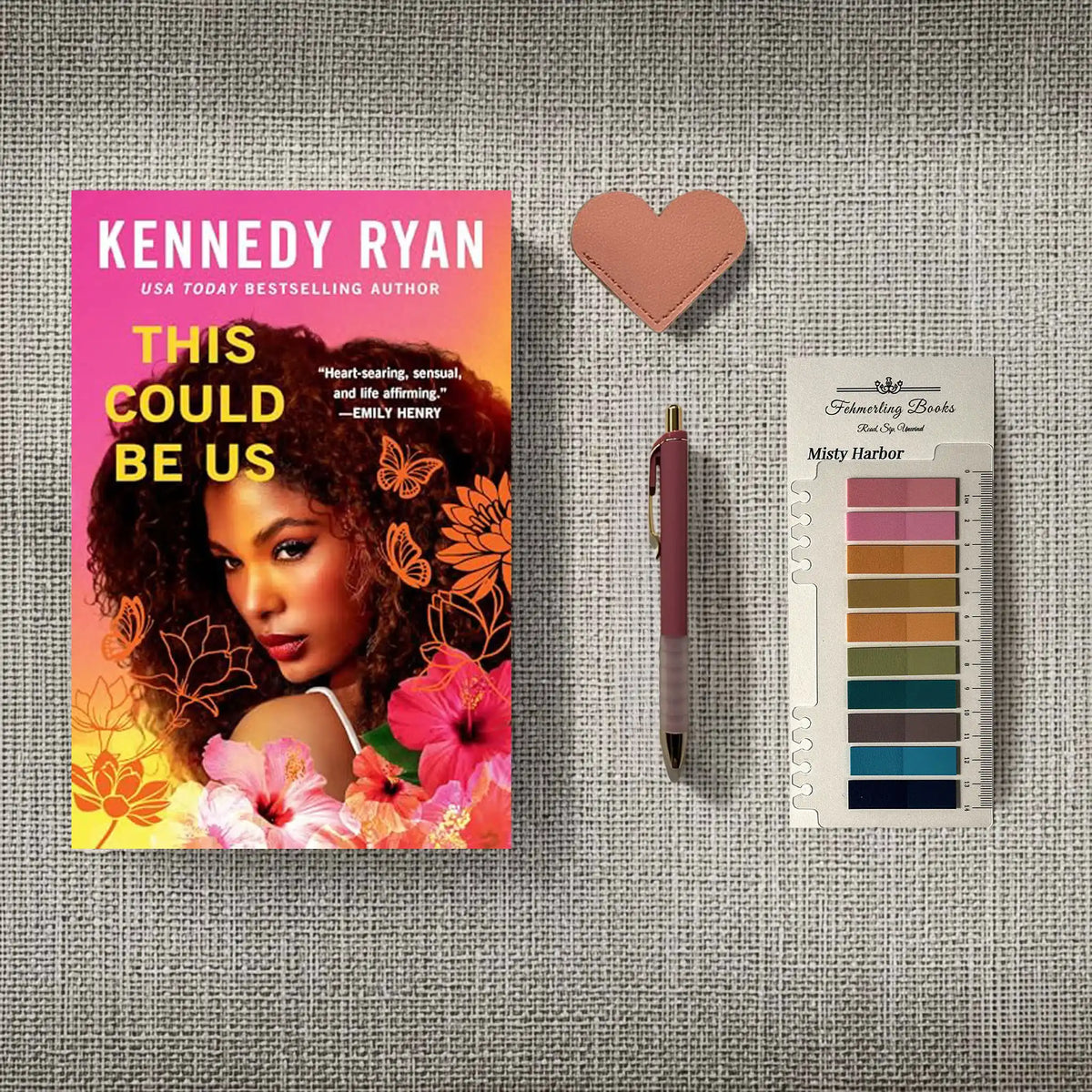 A styled flat-lay featuring the book 'This Could Be Us' by Kennedy Ryan, a pink heart-shaped leather bookmark, a pink pen, and a 'Fehmerling Books' Misty Harbor annotation tab card, arranged on a gray textured fabric background.