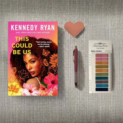A styled flat-lay featuring the book 'This Could Be Us' by Kennedy Ryan, a pink heart-shaped leather bookmark, a pink pen, and a 'Fehmerling Books' Misty Harbor annotation tab card, arranged on a gray textured fabric background.