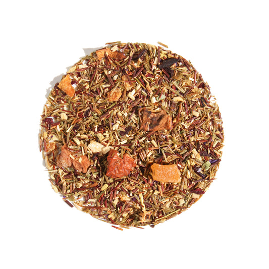 A close-up, overhead view of Plum Deluxe Dragonfruit Desire Herbal Tea arranged in a neat circle against a white background. The loose leaf blend features fine green rooibos needles mixed with visible chunks of dried mango and strawberry, citrus peels, and vibrant petals.