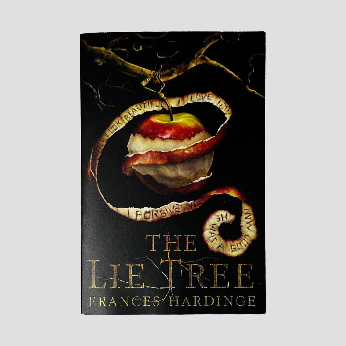 Front cover of The Lie Tree by Frances Hardinge in Like New paperback condition, featuring gold foil title lettering beneath a peeled apple hanging from a dark branch with inscribed text.