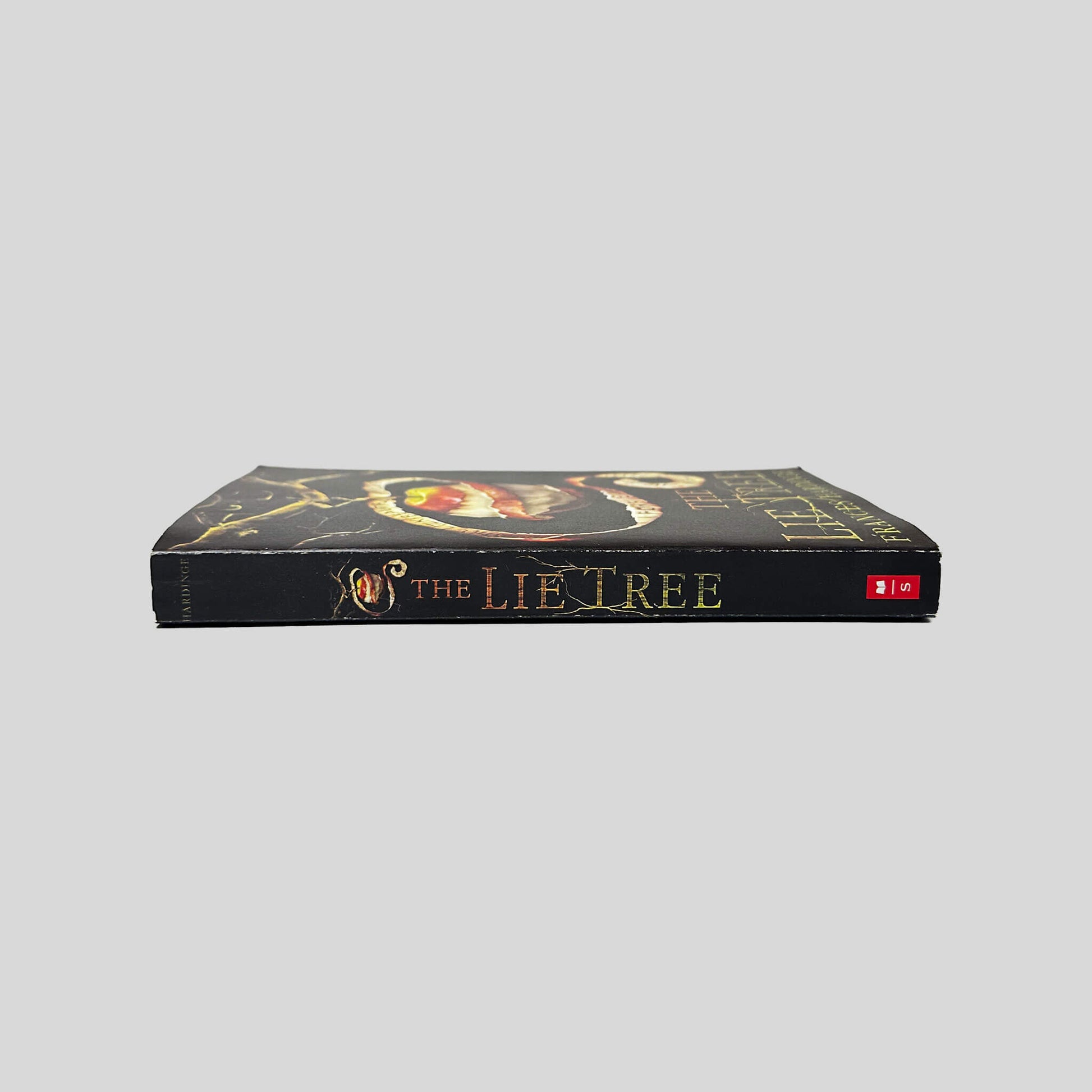 Unbroken spine view of The Lie Tree paperback by Frances Hardinge displaying gold foil title lettering, red Scholastic logo, crisp edges, and clean binding.