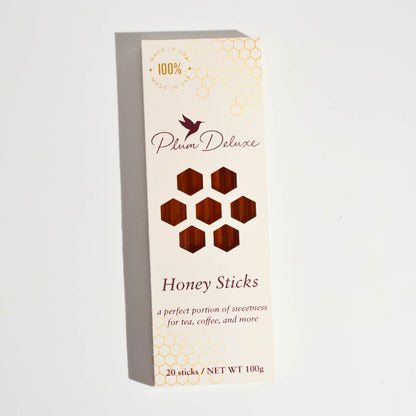 Product packaging for Plum Deluxe Honey Sticks. The box features the Plum Deluxe hummingbird logo, a honeycomb window displaying the amber honey, and text reading 'Honey Sticks: a perfect portion of sweetness for tea, coffee, and more'.