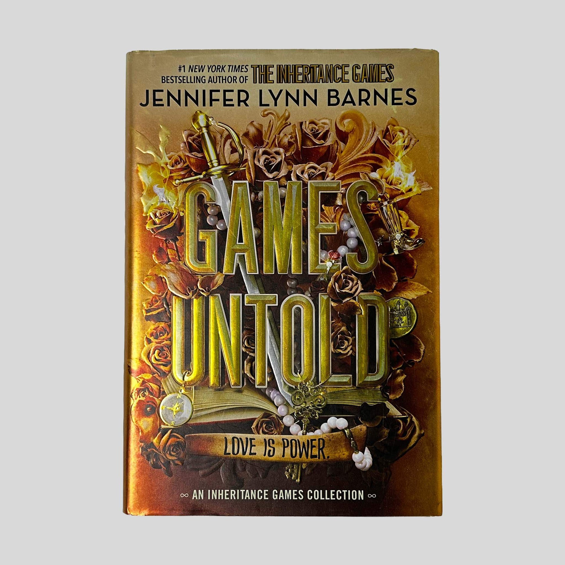 Front cover dust jacket of Games Untold by Jennifer Lynn Barnes in Like New hardcover condition, featuring gold metallic title lettering over warm amber and copper filigree art with roses, a sword, and pearl jewelry.