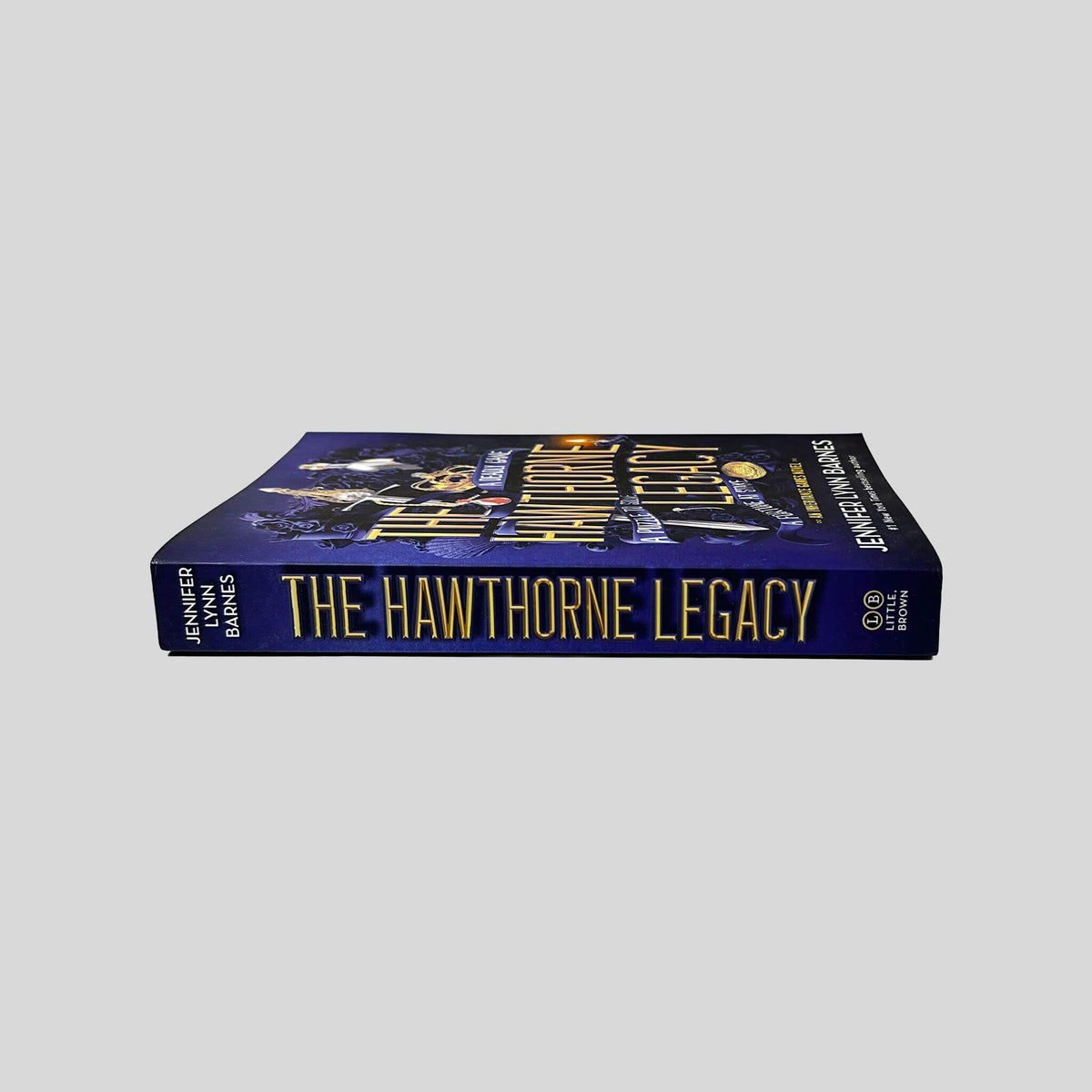 Unbroken spine view of The Hawthorne Legacy paperback by Jennifer Lynn Barnes displaying metallic gold title lettering, pristine edges, and clean binding.
