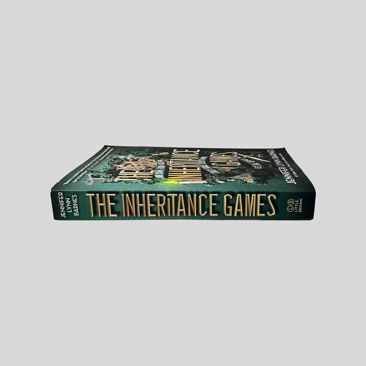 Unbroken spine view of The Inheritance Games paperback by Jennifer Lynn Barnes showing metallic gold title lettering, pristine edges, and clean binding.