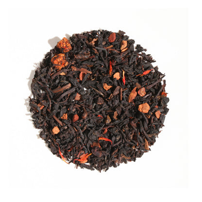A close-up, overhead view of Plum Deluxe Maple Pecan Holiday Tea arranged in a neat circle against a white background. The loose leaf blend features dark, rolled black and oolong tea leaves accented with chunky nut pieces, spices, and vivid red and orange petals.