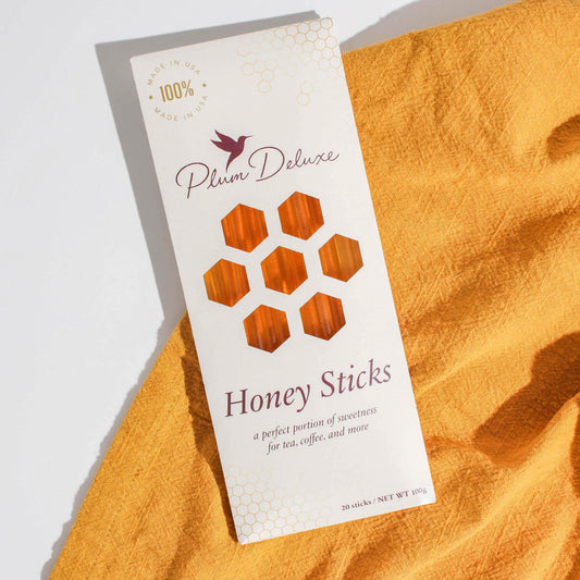 A lifestyle shot of Plum Deluxe Honey Sticks packaging lying on a warm yellow fabric. The box displays the Plum Deluxe logo, reads '100% Made in USA', and has peek-through honeycomb windows showing the golden honey.
