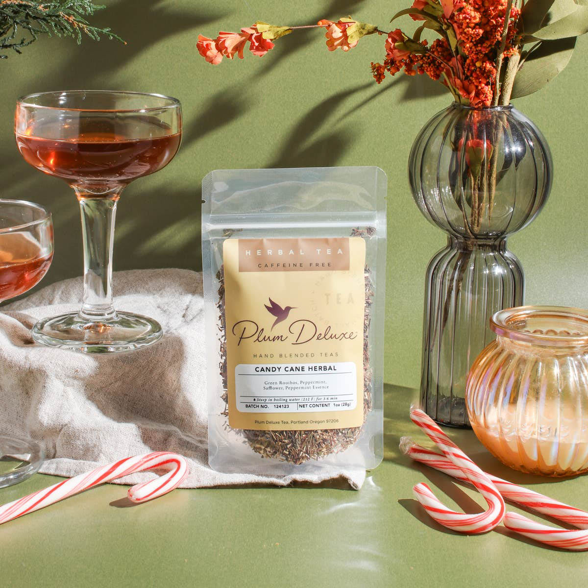 A clear pouch of Plum Deluxe Candy Cane Herbal Tea rests on a beige cloth against an olive green background. Surrounding the tea are three classic red and white striped candy canes, two glass coupes filled with steeped tea, a glowing glass candle, and a vase of orange winter florals.