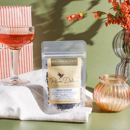 A clear pouch of Plum Deluxe Eggnog Oolong Tea rests on a beige cloth against an olive green background. Next to the tea is a glass coupe filled with amber tea, a red and white striped wrapped gift box, a ribbed glass candle, and a vase of festive orange florals.