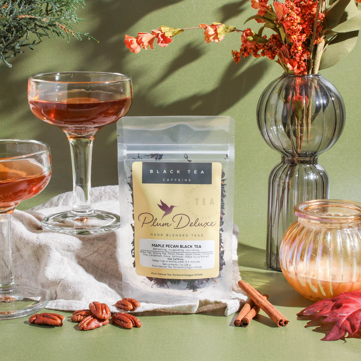 A clear pouch of Plum Deluxe Maple Pecan Holiday Black Tea rests on a beige linen cloth against an olive green background. It is surrounded by two elegant glass coupes filled with brewed amber tea, scattered pecans, cinnamon sticks, a glowing glass candle, and a vase of autumn foliage.