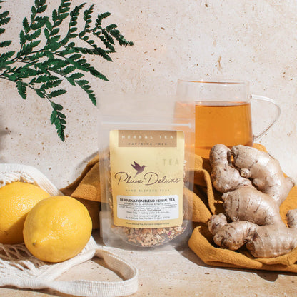 A lifestyle image featuring a bag of Plum Deluxe Rejuvenation Blend Herbal Tea. The tea is displayed on a textured countertop next to a glass mug of hot tea, fresh ginger root, lemons in a string bag, and a green botanical frond.