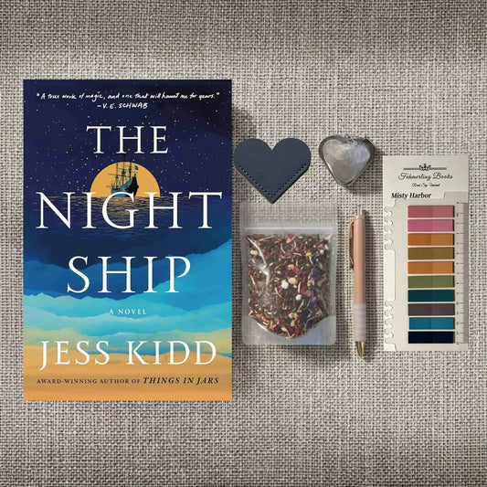 Book cover for 'The Night Ship' by Jess Kidd is displayed next to a flat-lay arrangement on a textured beige fabric background, including a heart-shaped leather bookmark and tea infuser, a small bag of herbal tea, a pen, and Misty Harbor annotation tabs.