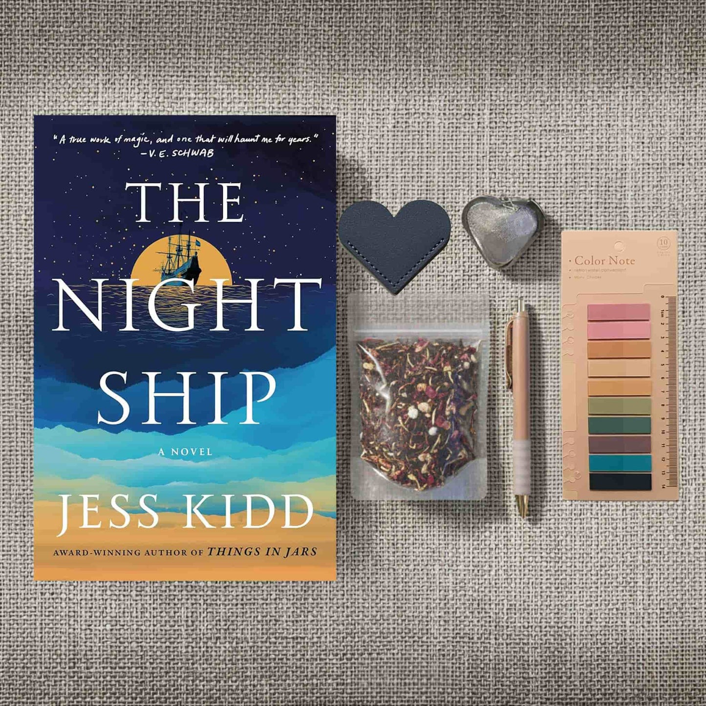 A product image for the "Reader Retreat Bundle" showcasing the New condition paperback of "The Night Ship" alongside expanded reading accessories. In addition to the heart-shaped bookmark, muted annotation tabs, and a rose-gold pen, this set includes a heart-shaped metal tea infuser and a clear packet of floral loose-leaf tea.