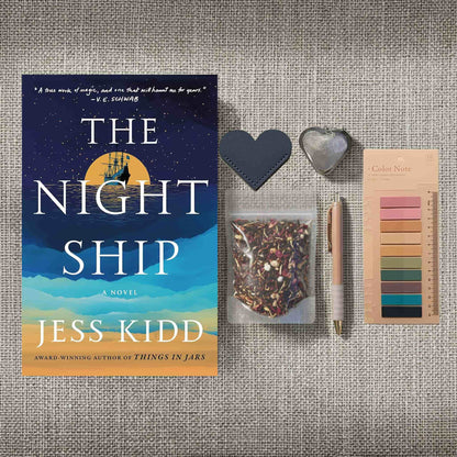 A product image for the "Reader Retreat Bundle" showcasing the New condition paperback of "The Night Ship" alongside expanded reading accessories. In addition to the heart-shaped bookmark, muted annotation tabs, and a rose-gold pen, this set includes a heart-shaped metal tea infuser and a clear packet of floral loose-leaf tea.
