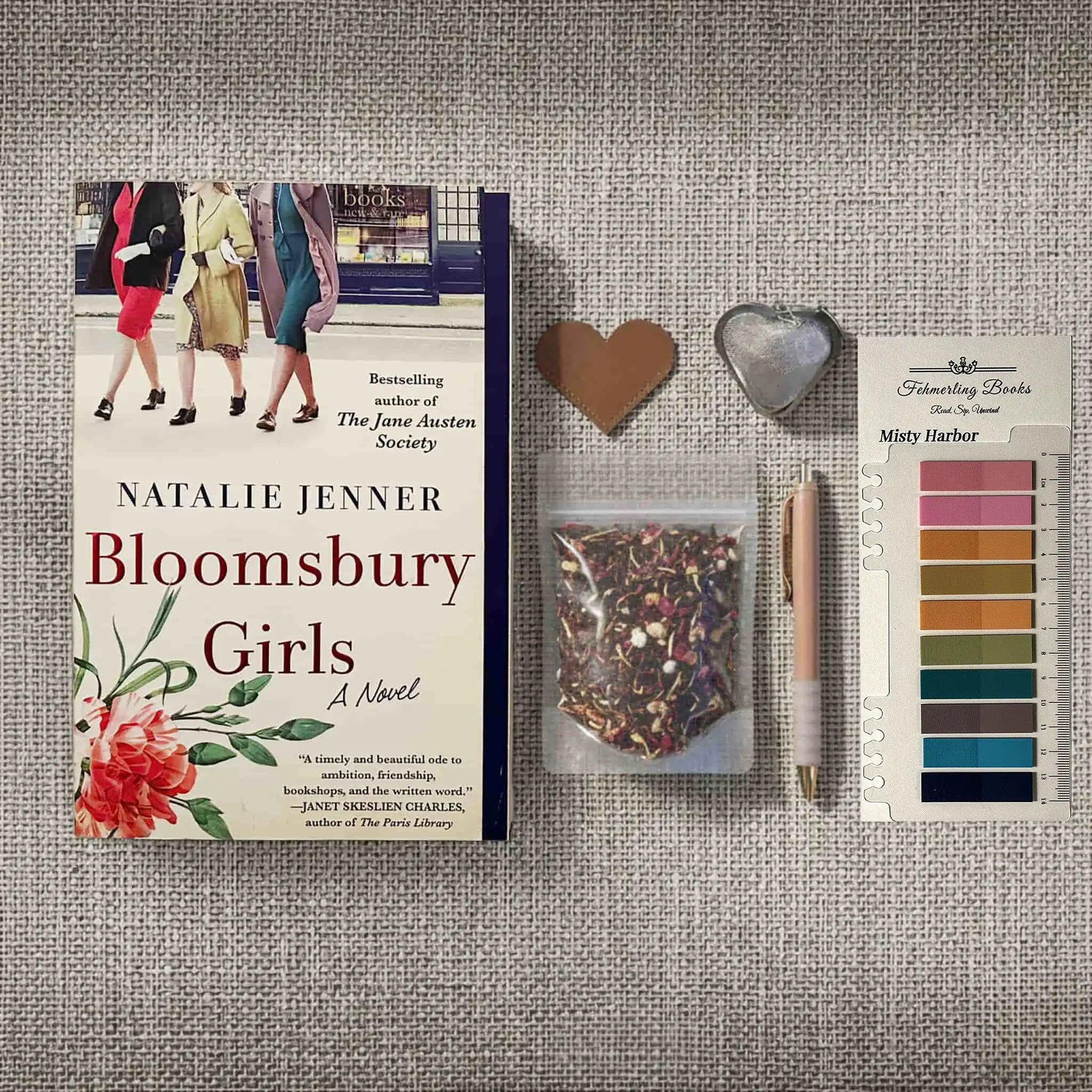 A flat-lay arrangement featuring the book 'Bloomsbury Girls' by Natalie Jenner, accompanied by a heart-shaped leather bookmark and tea infuser, Misty Harbor annotation tabs from Fehmerling Books, a pen, and a small bag of herbal tea on a textured gray fabric background.