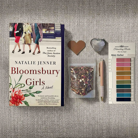 A flat-lay arrangement featuring the book 'Bloomsbury Girls' by Natalie Jenner, accompanied by a heart-shaped leather bookmark and tea infuser, Misty Harbor annotation tabs from Fehmerling Books, a pen, and a small bag of herbal tea on a textured gray fabric background.