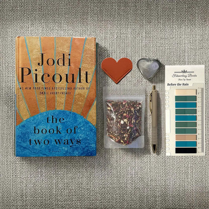 A styled flat-lay featuring the book 'The Book of Two Ways' by Jodi Picoult, accompanied by a small pouch of loose-leaf tea, a pen, a heart-shaped leather bookmark and tea infuser, and Before the Rain annotation tabs from Fehmerling Books, all arranged on a textured gray fabric background.