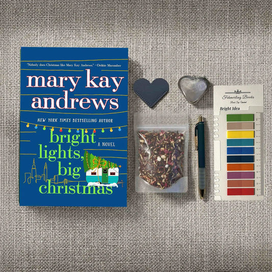 A styled flat-lay featuring the book 'Bright Lights, Big Christmas' by Mary Kay Andrews, accompanied by a heart-shaped tea infuser and leather bookmark, a clear bag of herbal tea, Bright Idea annotation tabs, and a pen, all arranged on a textured gray fabric background.