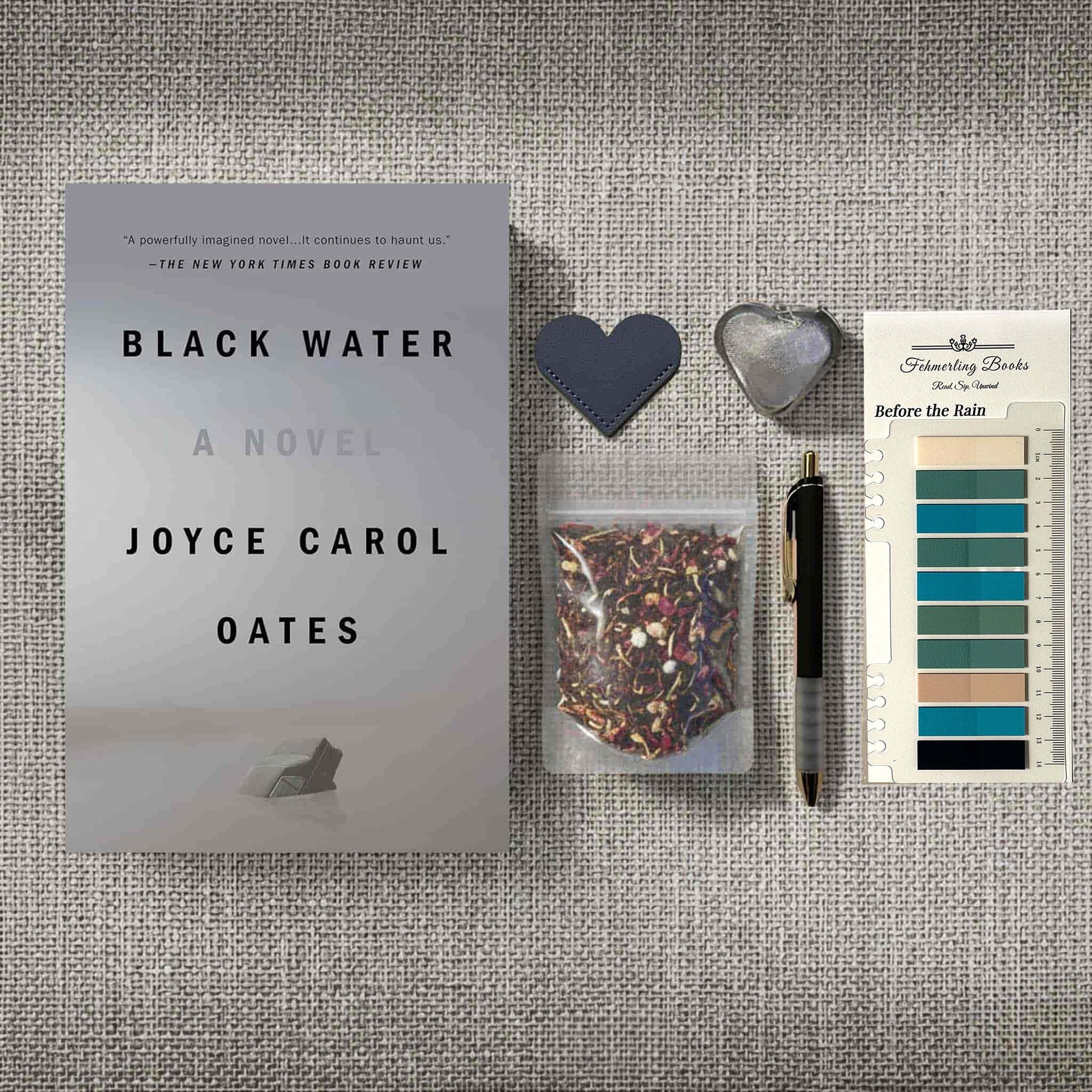 A styled flat-lay featuring the book 'Black Water' by Joyce Carol Oates alongside a small clear bag of tea, a black pen, a dark heart-shaped bookmark, a silver heart-shaped tea infuser, and annotation tabs on a textured gray fabric background.