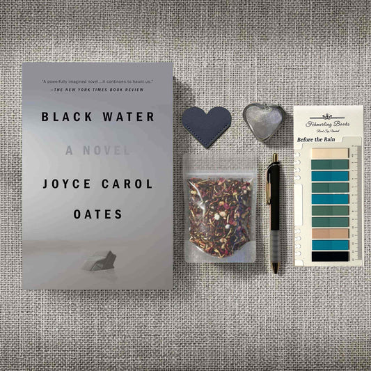 A styled flat-lay featuring the book 'Black Water' by Joyce Carol Oates alongside a small clear bag of tea, a black pen, a dark heart-shaped bookmark, a silver heart-shaped tea infuser, and annotation tabs on a textured gray fabric background.