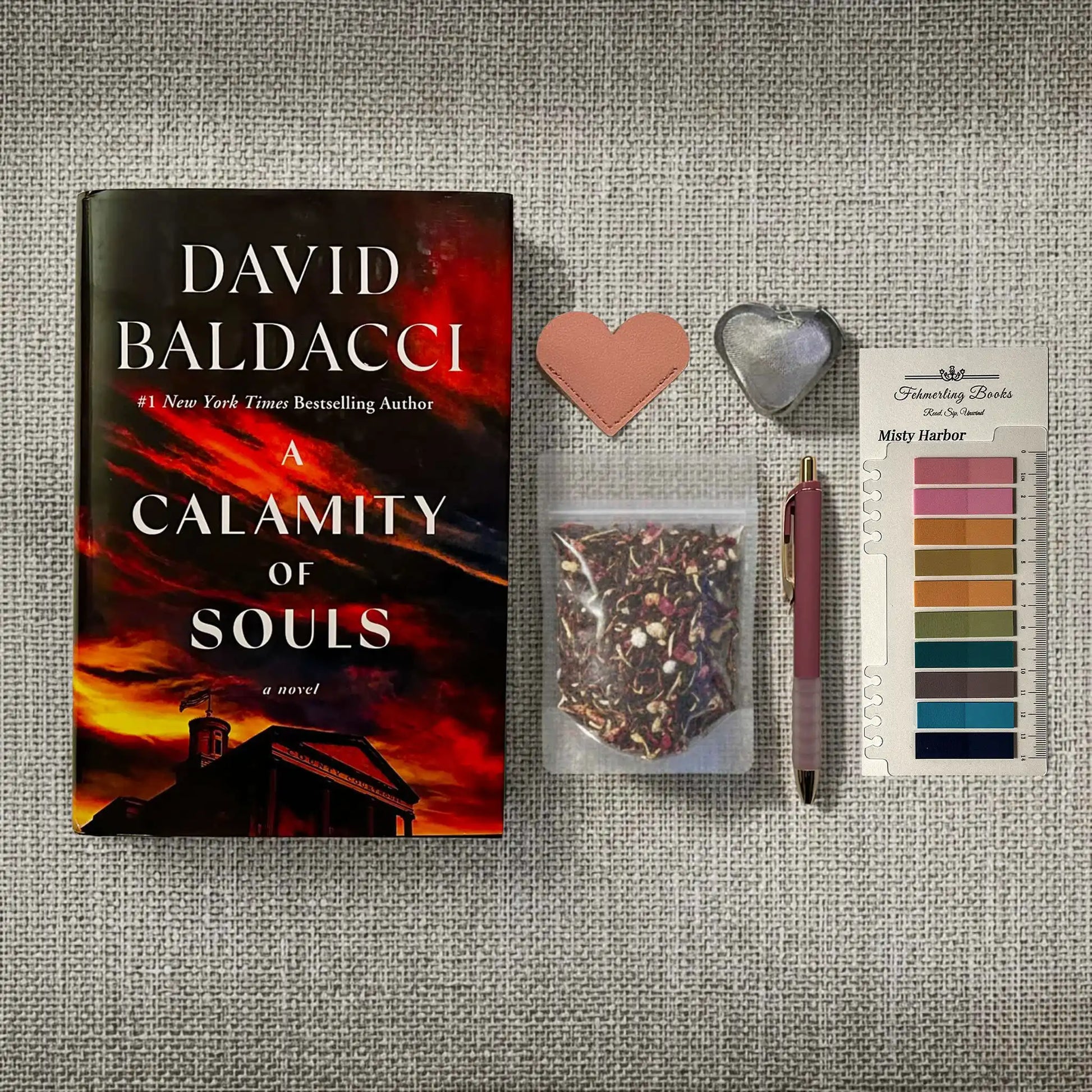 A styled flat-lay featuring the book 'A Calamity of Souls' by David Baldacci, alongside a pink heart-shaped leather bookmark and tea infuser, a small bag of loose-leaf tea, a pink pen, and Misty Harbor annotation tabs from Fehmerling Books on a textured gray fabric background.