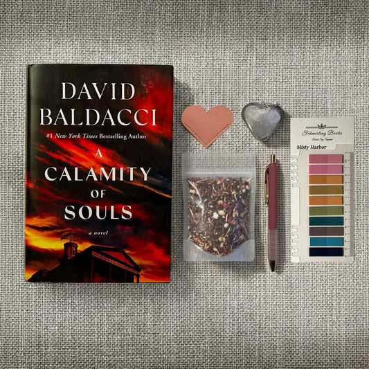 A styled flat-lay featuring the book 'A Calamity of Souls' by David Baldacci, alongside a pink heart-shaped leather bookmark and tea infuser, a small bag of loose-leaf tea, a pink pen, and Misty Harbor annotation tabs from Fehmerling Books on a textured gray fabric background.