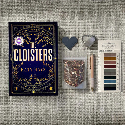 A styled flat-lay featuring the book 'The Cloisters' by Katy Hays, accompanied by a bag of herbal tea, Gothic Romance annotation tabs from Fehmerling Books, a pen, and heart-shaped leather bookmark and tea infuser on a textured gray fabric background.