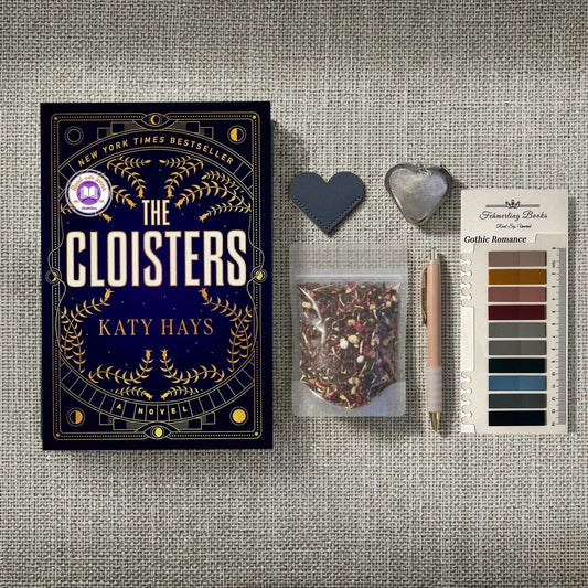 A styled flat-lay featuring the book 'The Cloisters' by Katy Hays, accompanied by a bag of herbal tea, Gothic Romance annotation tabs from Fehmerling Books, a pen, and heart-shaped leather bookmark and tea infuser on a textured gray fabric background.