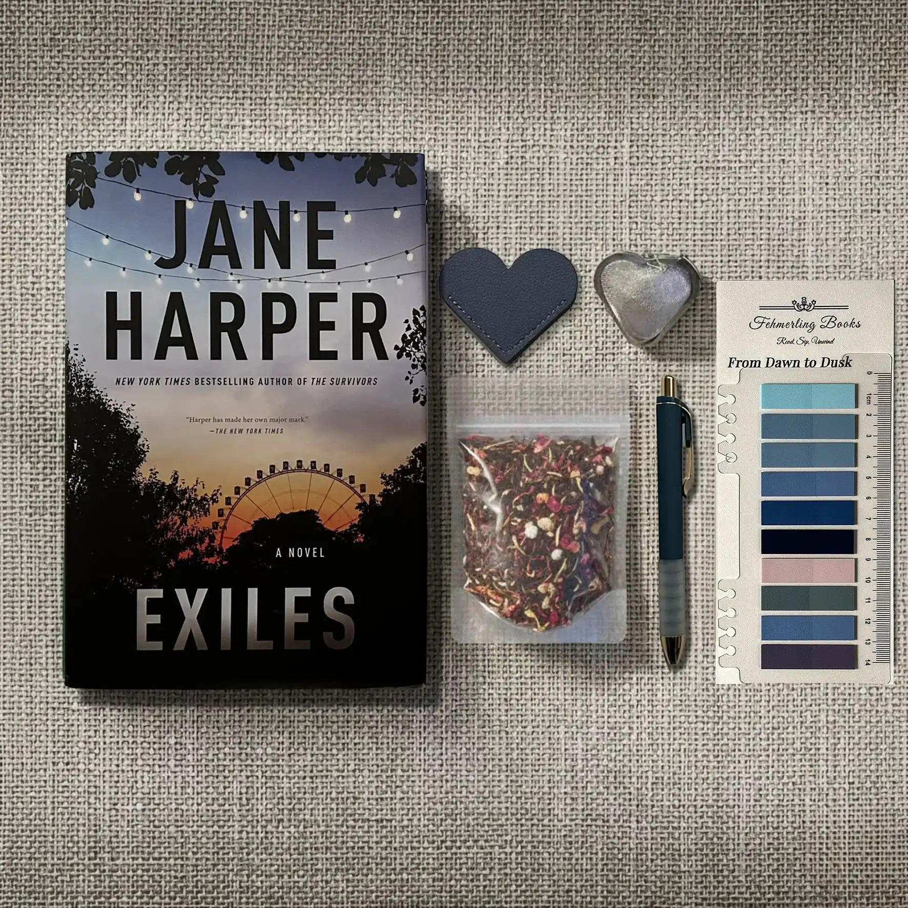 A styled flat-lay featuring Jane Harper's novel 'Exiles' on a textured linen background, accompanied by a small bag of herbal tea, a heart-shaped tea infuser and leather bookmark, a pen, and From Dawn to Dusk annotation tabs.