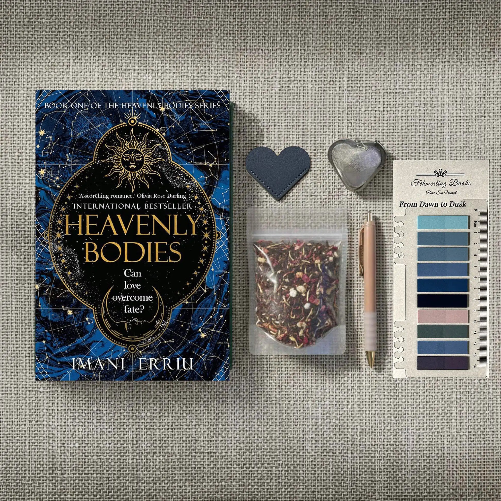 A styled flat-lay featuring the book 'Heavenly Bodies' by Imani Erriu, accompanied by a notebook with From Dawn to Dusk annotation tabs, a pen, a small bag of herbal tea, a heart-shaped tea infuser and leather bookmark on a textured gray fabric background.