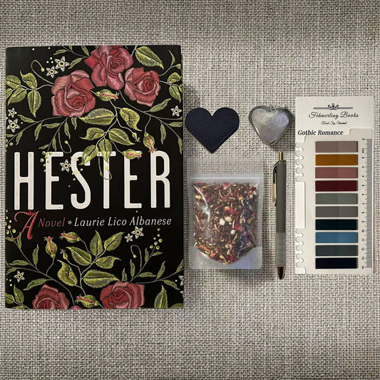 A styled flat-lay of a book titled 'Hester' by Laurie Lico Albanese, displayed alongside a small heart-shaped leather bookmark and tea infuser, a packet of herbal tea and Gothic Romance annotation tabs from Fehmerling Books. The book cover features a black background decorated with pink roses and green leaves.