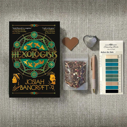 A styled flat-lay of a book titled 'The Hexologists' by Josiah Bancroft, arranged on a textured gray fabric background alongside a small bag of loose herbal tea, a rose gold pen, a heart-shaped leather bookmark and tea infuser, and Before the Rain annotation tabs from Fehmerling Books.
