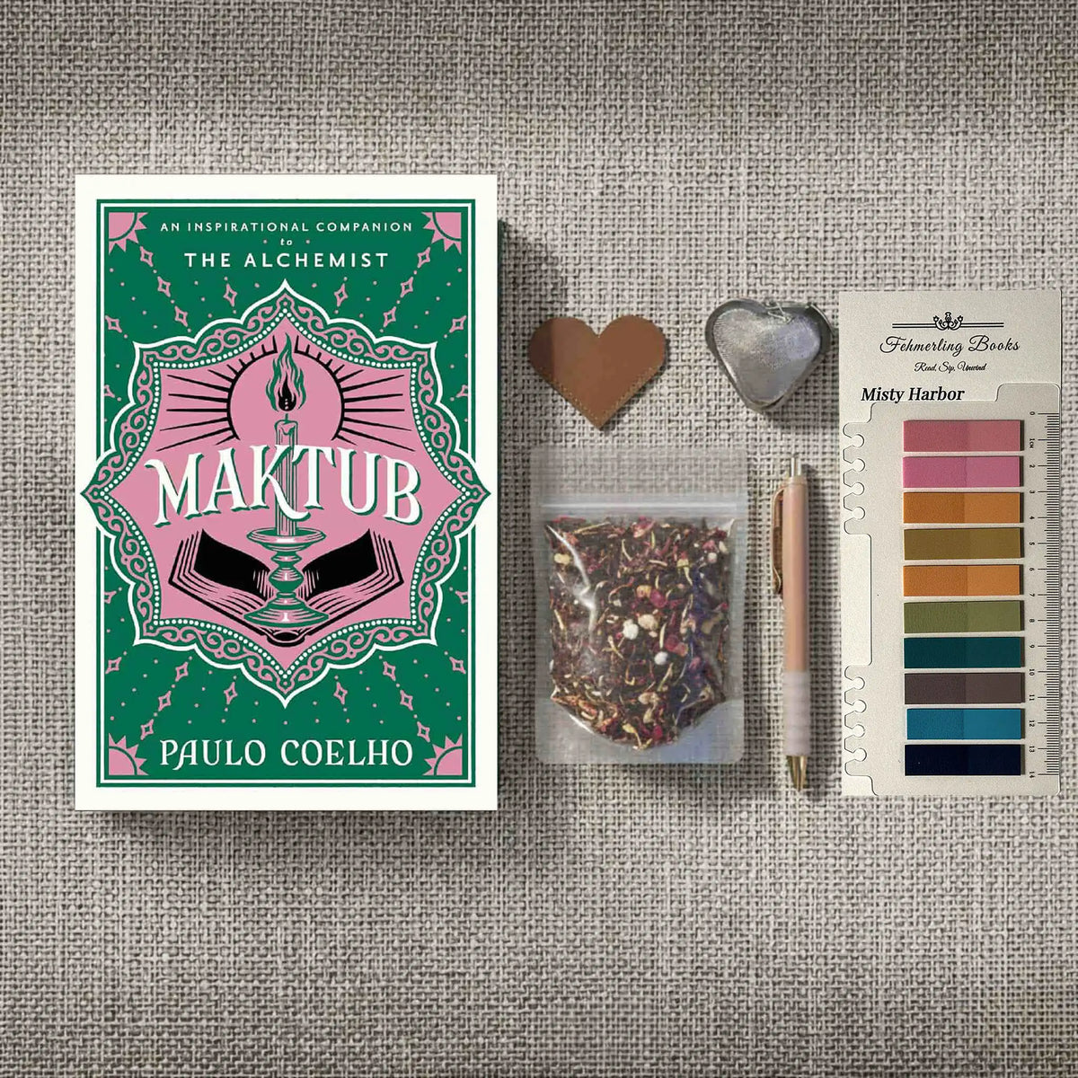 A styled flat-lay featuring the book 'Maktub' by Paulo Coelho on the left, alongside a small bag of herbal tea, a rose gold pen, a heart-shaped leather bookmark and tea infuser, and Misty Harbor annotation tabs from Fehmerling Books on the right, all arranged on a textured gray fabric background.