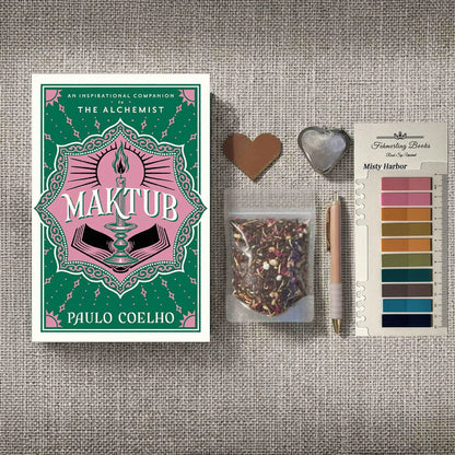 A styled flat-lay featuring the book 'Maktub' by Paulo Coelho on the left, alongside a small bag of herbal tea, a rose gold pen, a heart-shaped leather bookmark and tea infuser, and Misty Harbor annotation tabs from Fehmerling Books on the right, all arranged on a textured gray fabric background.