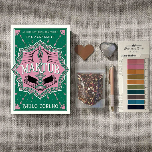 A styled flat-lay featuring the book 'Maktub' by Paulo Coelho on the left, alongside a small bag of herbal tea, a rose gold pen, a heart-shaped leather bookmark and tea infuser, and Misty Harbor annotation tabs from Fehmerling Books on the right, all arranged on a textured gray fabric background.