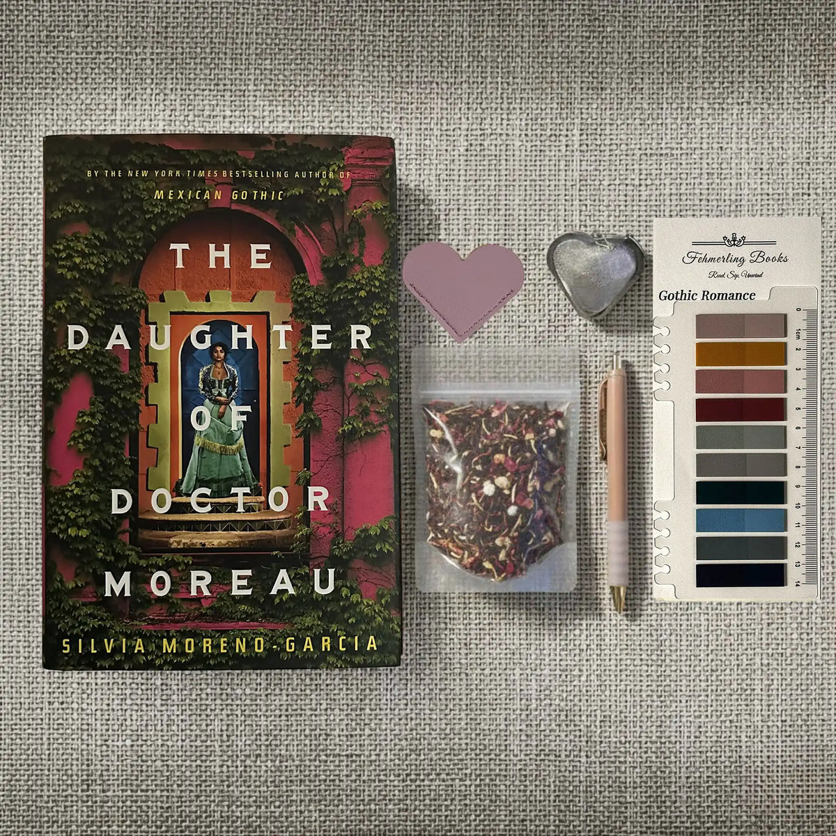 A styled flat-lay featuring the book 'The Daughter of Doctor Moreau' by Silvia Moreno-Garcia, accompanied by a heart-shaped leather bookmark and tea infuser, a bag of herbal tea, a rose gold pen, and Gothic Romance annotation tabs from Fehmerling Books on a textured beige fabric background.