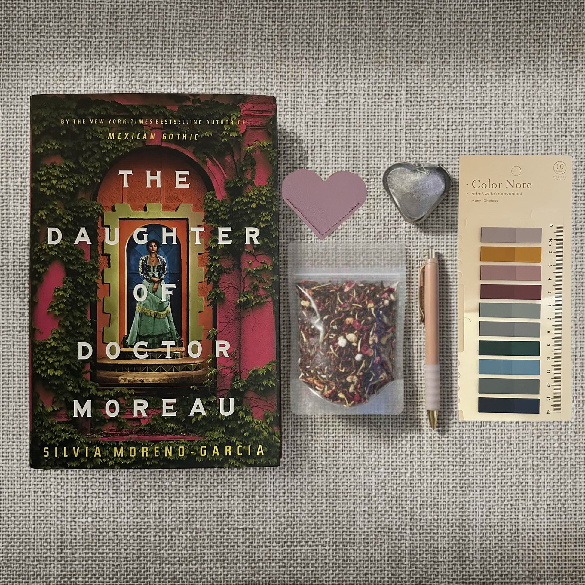 Reader's Retreat bundle flat lay for The Daughter of Doctor Moreau by Silvia Moreno-Garcia. Includes the lush hardcover book, a brown Storyteller's journal with compass charm, heart-shaped tea infuser, artisanal Evening in the Garden tea, lavender heart bookmark, and Gothic Romance annotation tabs on a textured beige surface. Captures the premium tropical gothic aesthetic of this Fehmerling Books collection.