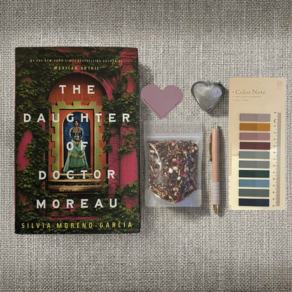 Reader's Retreat bundle flat lay for The Daughter of Doctor Moreau by Silvia Moreno-Garcia. Includes the lush hardcover book, a brown Storyteller's journal with compass charm, heart-shaped tea infuser, artisanal Evening in the Garden tea, lavender heart bookmark, and Gothic Romance annotation tabs on a textured beige surface. Captures the premium tropical gothic aesthetic of this Fehmerling Books collection.