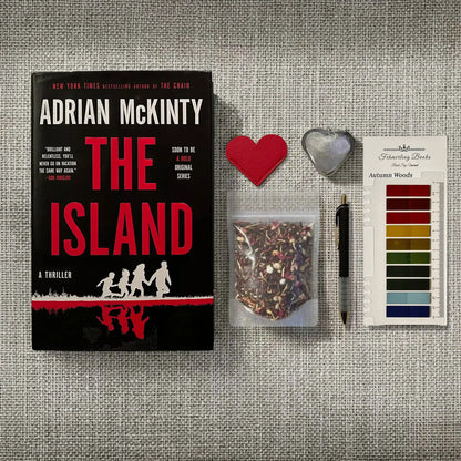 Book cover for 'The Island' by Adrian McKinty displayed on a textured gray fabric background, alongside a heat-shaped leather bookmark and tea infuser, a small bag of loose-leaf tea, a black pen, and Autumn Woods annotation tabs from Fehmerling Books.