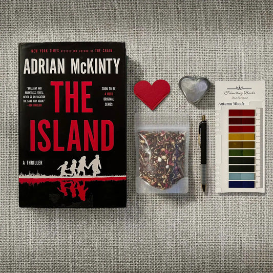 Book cover for 'The Island' by Adrian McKinty displayed on a textured gray fabric background, alongside a heat-shaped leather bookmark and tea infuser, a small bag of loose-leaf tea, a black pen, and Autumn Woods annotation tabs from Fehmerling Books.