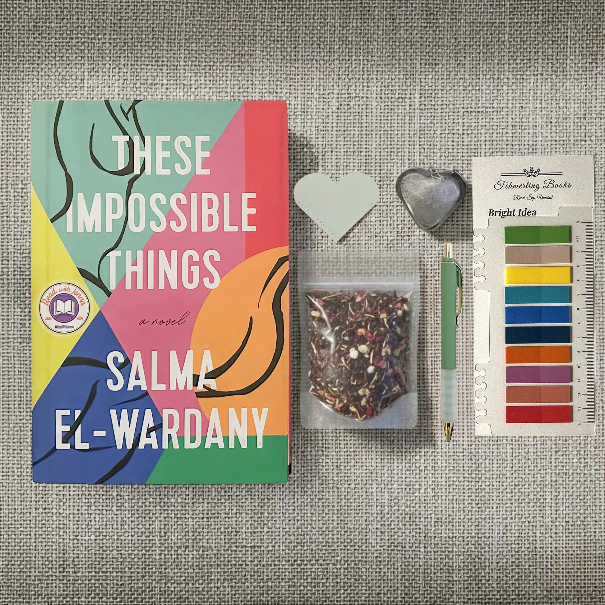 A styled flat-lay featuring the book 'These Impossible Things' by Salma El-Wardany, accompanied by a small clear bag of loose-leaf tea, a pastel green pen, heart-shaped tea infuser and leather bookmark, and Bright Idea annotation tabs, all arranged on a textured gray linen background.