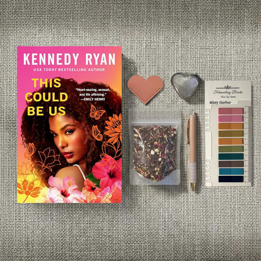A styled flat-lay featuring the book 'This Could Be Us' by Kennedy Ryan, accompanied by a heart-shaped leather bookmark and tea infuser, a clear bag of herbal tea, a rose gold pen, and a 'Fehmerling Books' Misty Harbor annotation tab card on a textured gray fabric background.
