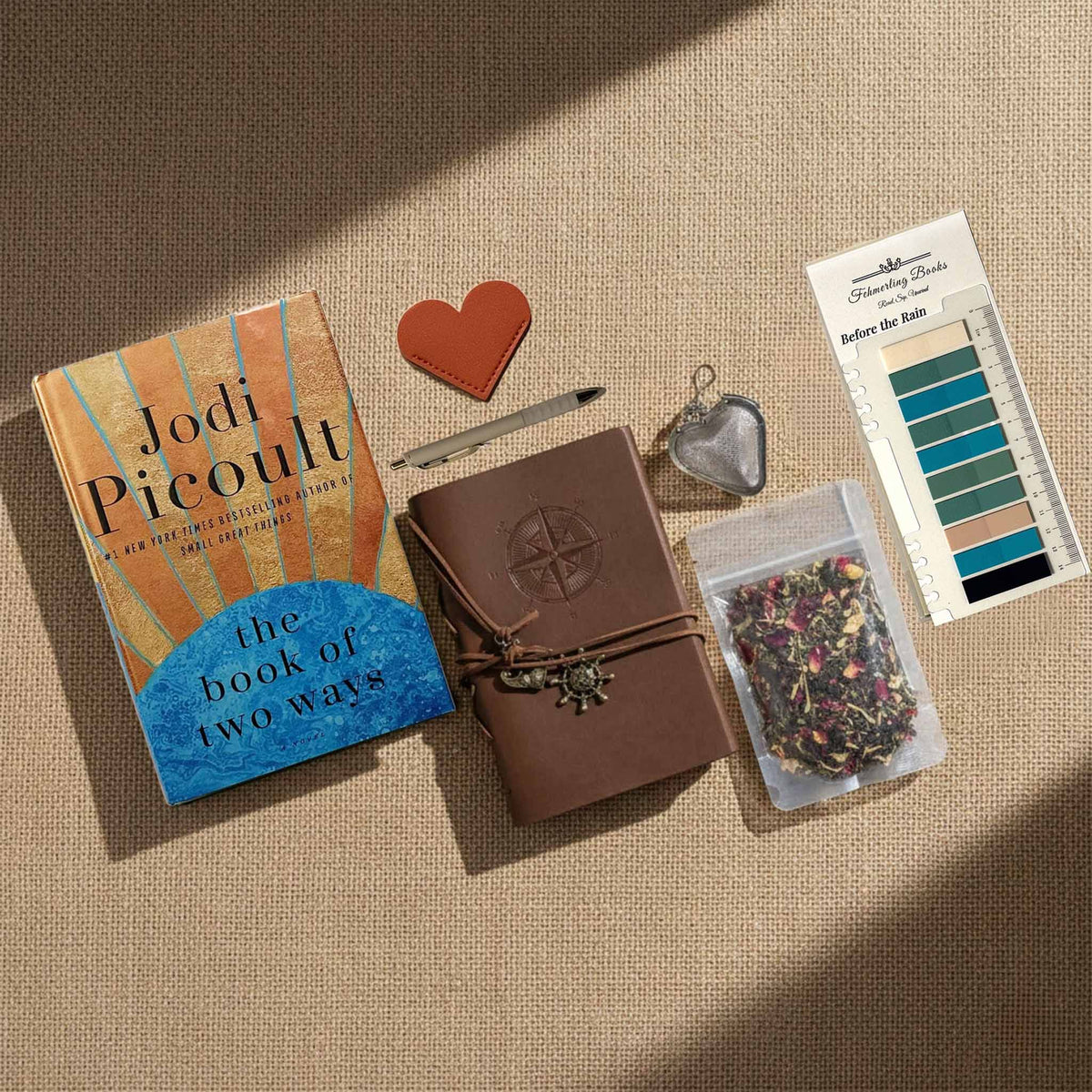 A styled flat-lay arrangement on a beige fabric background featuring The Book of Two Ways by Jodi Picoult, a brown leather journal with an embossed compass, a heart-shaped leather bookmark and tea infuser, a tea bag, and Before the Rain annotation tabs from Fehmerling Books.