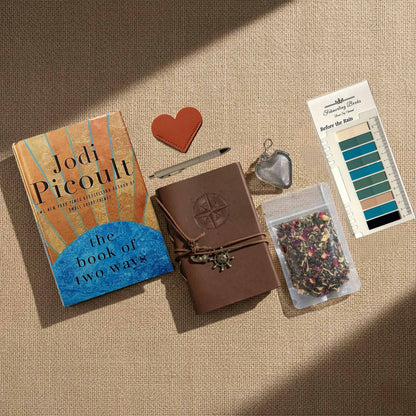 A styled flat-lay arrangement on a beige fabric background featuring The Book of Two Ways by Jodi Picoult, a brown leather journal with an embossed compass, a heart-shaped leather bookmark and tea infuser, a tea bag, and Before the Rain annotation tabs from Fehmerling Books.