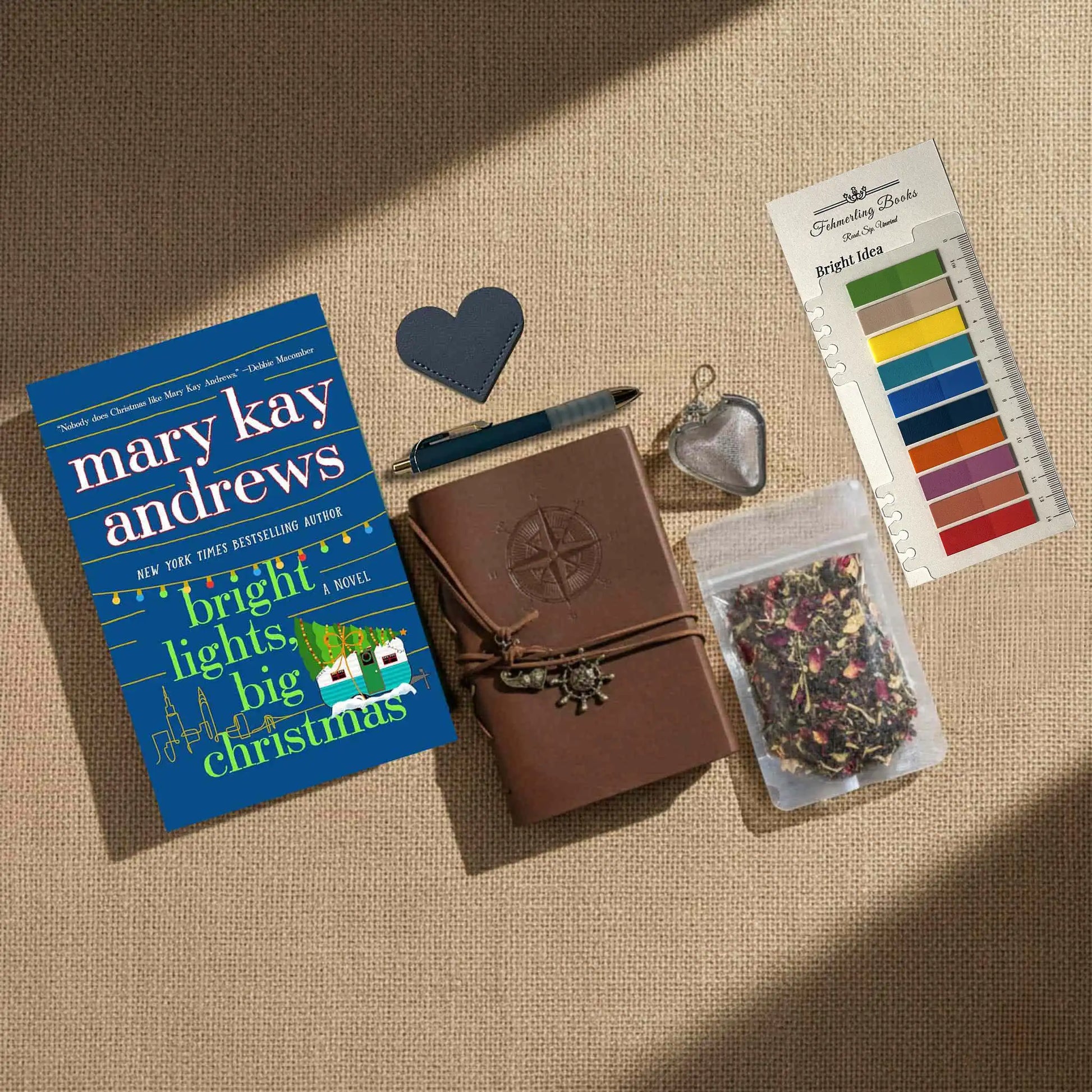 A styled flat-lay of a book gift set on a beige burlap background. The set includes a copy of the book 'Bright Lights, Big Christmas' by Mary Kay Andrews, a brown leather journal with a compass emblem, a blue pen, a heart-shaped leather bookmark, a metal tea infuser, a packet of dried flower tea, and Bright Idea annotation tabs.