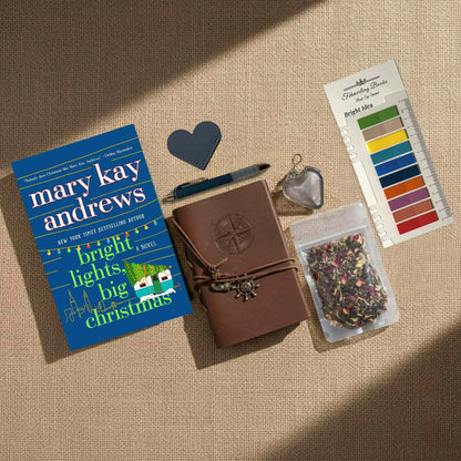 A styled flat-lay of a book gift set on a beige burlap background. The set includes a copy of the book 'Bright Lights, Big Christmas' by Mary Kay Andrews, a brown leather journal with a compass emblem, a blue pen, a heart-shaped leather bookmark, a metal tea infuser, a packet of dried flower tea, and Bright Idea annotation tabs.