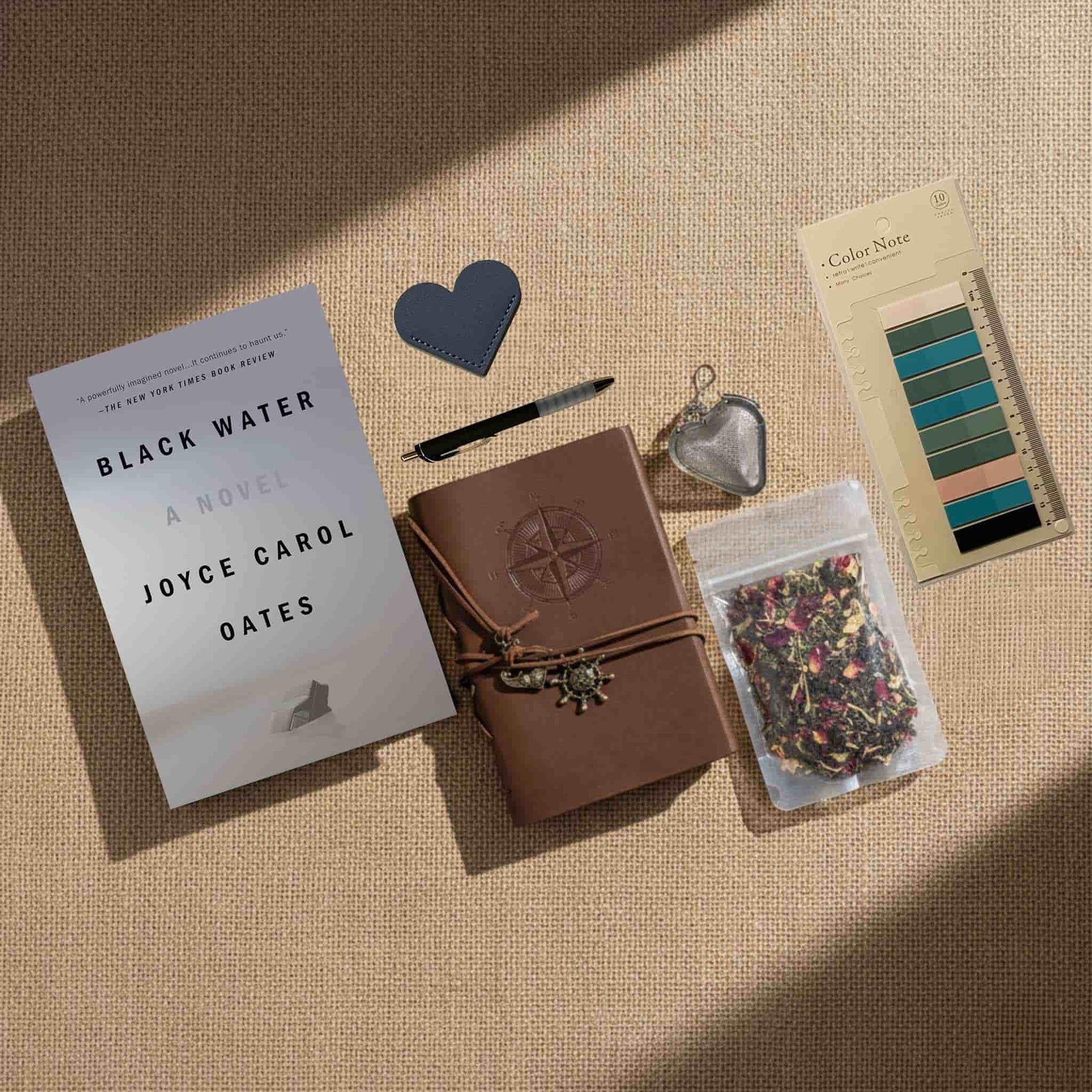 Dark academia aesthetic gift bundle with 'Black Water' by Joyce Carol Oates, a heart-shaped leather bookmark, and color palette swatches on a neutral textured background.