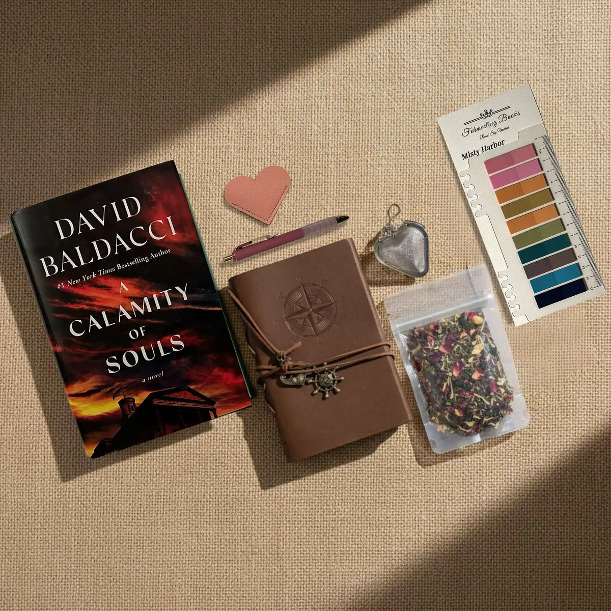 A styled flat-lay arrangement on a beige fabric background featuring a book by David Baldacci, a brown leather journal with a wrap closure, a pink heart-shaped leather book mark and tea infuser, Misty Harbor annotation tabs, and a small bag of dried flower tea.